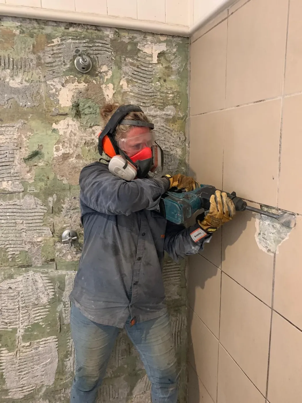 Stacey removing wall tiles during a bathroom renovation, wearing protective equipment.